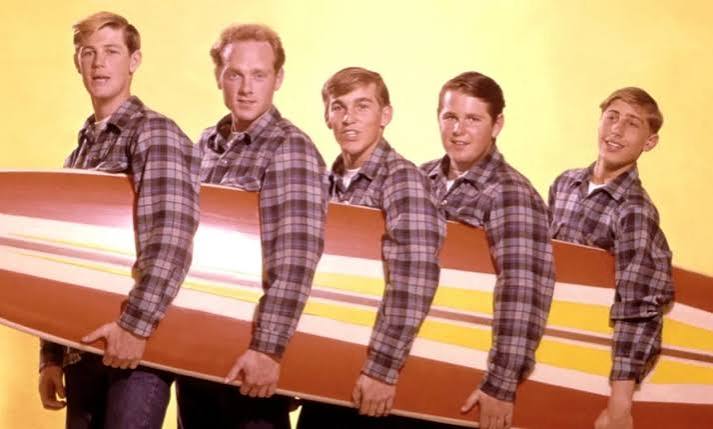 Cinco jóvenes con camisas a cuadros posan junto a una tabla de surf colorida sobre fondo amarillo, The Beach Boys